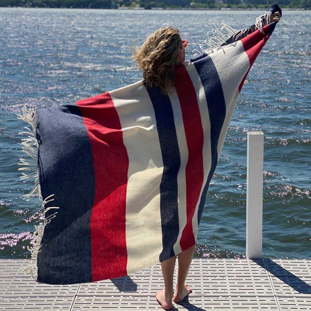 Person on a lake-side dock wearing BIG LOViE Peace collection Freedom blanket with fringe blowing in the wind.
