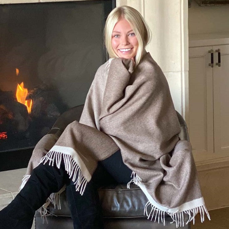 Smiling woman wrapped in BIG LOViE Soul Coco 100 percent baby alpaca blanket sitting in front of fireplace.