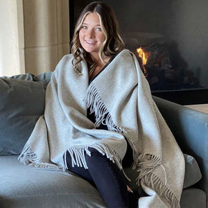Smiling woman wrapped in BIG LOViE Soul KJ 100% Alpaca Eco blanket in Silver sitting on couch in front of fireplace.