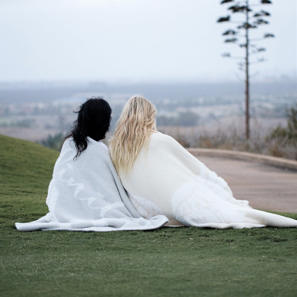 Two woman sitting together on grass, wrapped in Love and Wings Blanket from the Dream Collection, micro-chenille and feather yarn