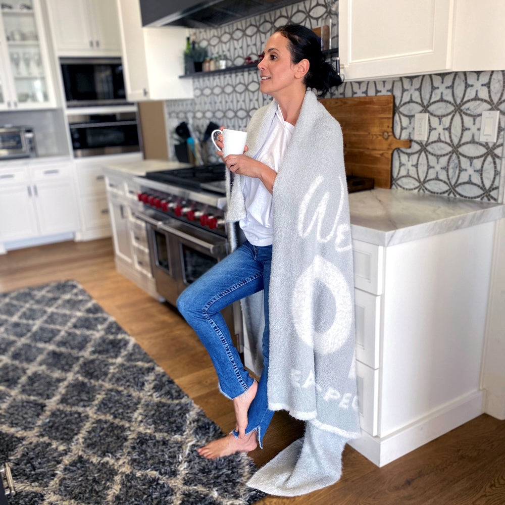 Woman drinking coffee in kitchen, One Blanket from Dream Collection draped over her, decorative rug, white cabinets