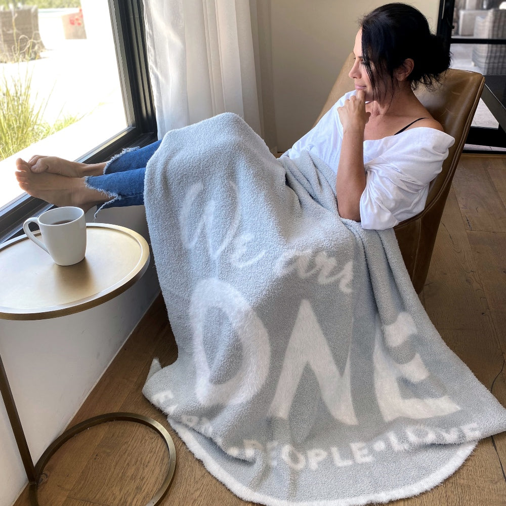 Woman sitting on chair with feet up, drinking coffee, wrapped in the One Blanket from Dream Collection, micro-chenille and feather yarn, light grey