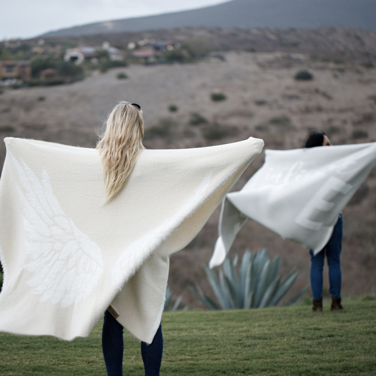 Woman spreading their wings in the Wings Blanket from the Dream Collection, micro-chenille and feather yarn, mountains in distance