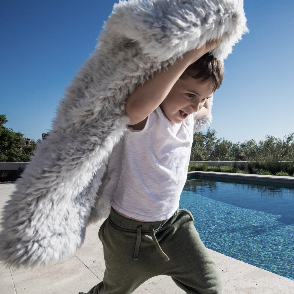 Young boy running with BIG LOViE Little Guardian Angel Blanket in Silver Cloud over his head, pool and trees in background